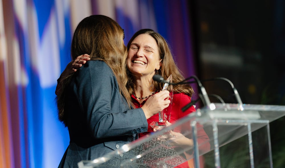 A woman in a red dress smiles and hugs a colleague on stage while receiving the AVID Global Achievement Award.
